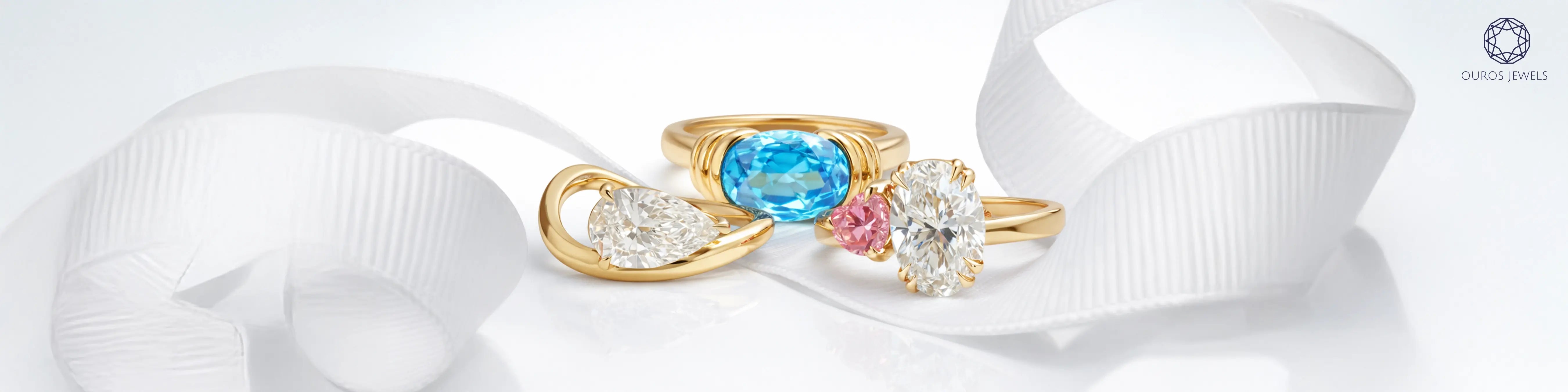 Three unique engagement rings in yellow gold featuring oval blue topaz east-west ring, oval diamond with pink diamond toi et moi ring and pear diamond wave ring by Ouros Jewels
