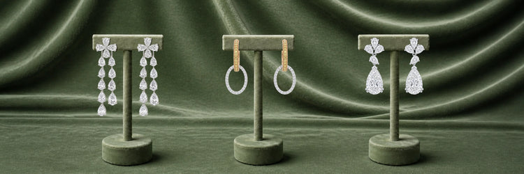 Three pairs of lab-grown diamond earrings on olive stands — white gold pear cut cascading chandelier drop earrings, yellow gold pavé oval hoop earrings, and white gold triple pear cluster with large pear drop earrings on green satin