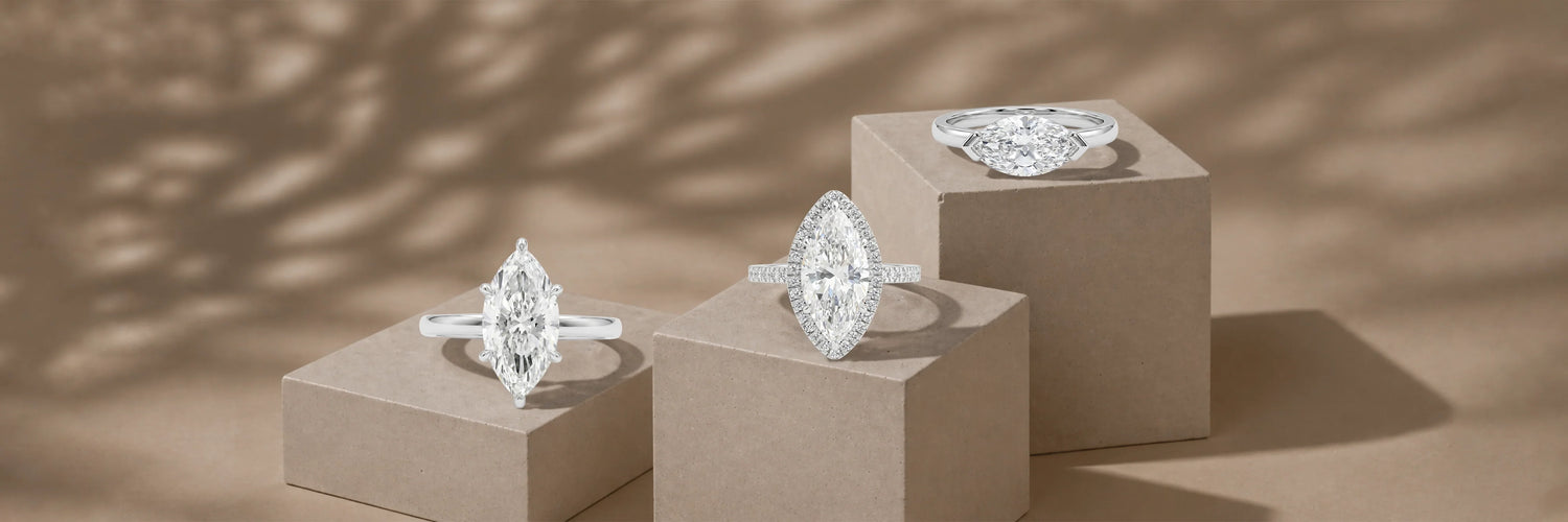 Three marquise cut lab-grown diamond engagement rings in white gold — classic six-prong solitaire, marquise diamond halo with pavé band, and east-west bezel solitaire on taupe pedestals with dappled light