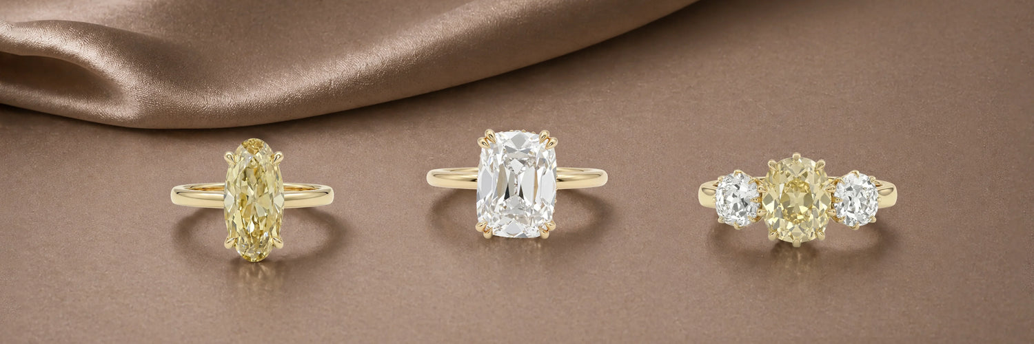 Three old cut lab-grown diamond engagement rings in yellow gold — fancy yellow oval solitaire, large old mine cushion cut solitaire with claw prongs, and fancy yellow three-stone ring with round white diamond side stones on champagne satin