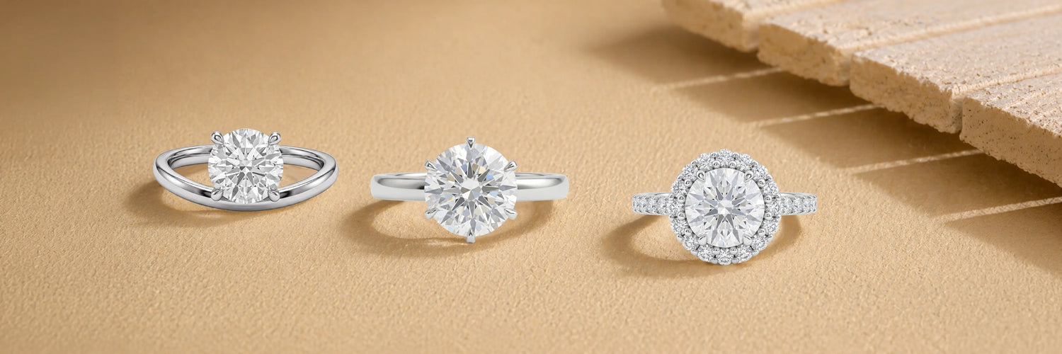 Three round cut lab-grown diamond engagement rings in white gold — bypass solitaire, six-prong classic solitaire, and round brilliant halo with pavé band on sandy beige surface