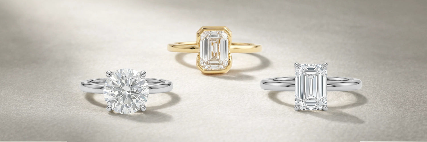 Three solitaire lab-grown diamond engagement rings displayed on a neutral surface — round brilliant cut in white gold, emerald cut bezel setting in yellow gold, and emerald cut four-prong solitaire in white gold