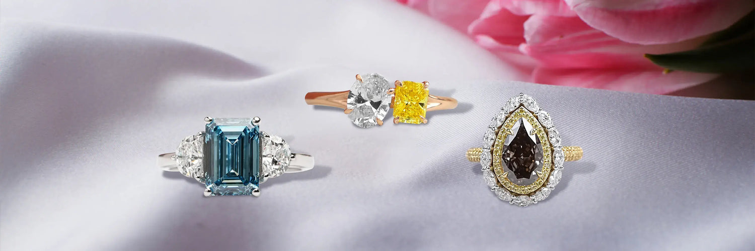 Four fancy colored lab-grown diamond rings in white gold — pink marquise halo, pink oval double halo with baguette shoulders, blue asscher cut solitaire, and fancy yellow pear cut solitaire on burgundy display