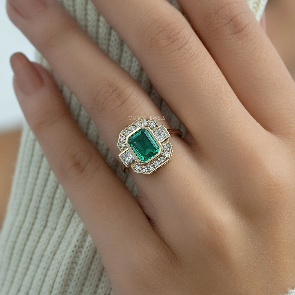 Three stone green emerald cut ring with bezel and halo diamond settings worn on a finger