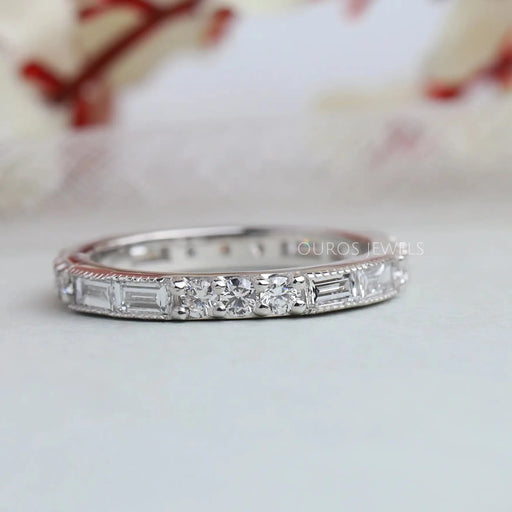 Baguette And Round Diamond Eternity Wedding Band is on soft white surface with blurred flora background.