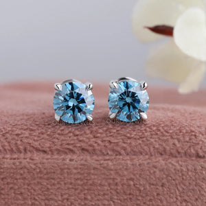 Blue Round Diamond Stud Earrings displayed on pink velvet box with blurred floral background.