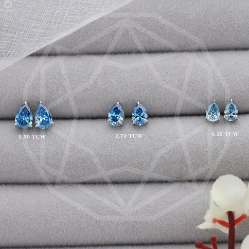 Blue Pear Shaped Diamond Stud Earrings: Three pairs of blue diamond stud earrings, each with different carat weights (0.90 TCW, 0.70 TCW, 0.50 TCW), displayed on a soft gray fabric background with a small white flower on the side.